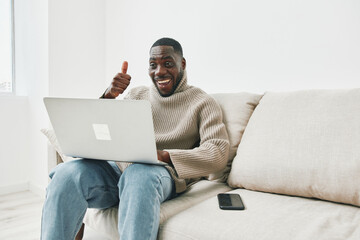 Happy young Black man working on a laptop at home, wearing a cozy sweater and showing a thumbs up while sitting on a light colored sofa