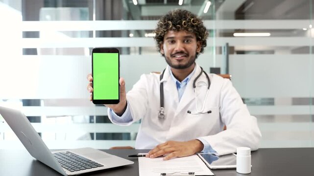 Young doctor showing mobile phone with green screen while sitting in hospital office. Color key, vertical template layout for advertising. Medical worker physician looking at camera holding smartphone