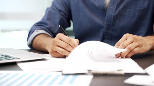 Close up of a male's hand signing documents at a desk at a workplace in a modern business office. The boss or manager looks through the folder with documentation and signs the contract with a pen