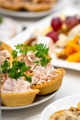 Close-up of savory tartlets with creamy pink filling and parsley garnish, served on a white plate at a buffet with assorted appetizers in the background