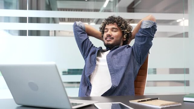 Happy businessman finished work on laptop while sitting at desk at workplace in a modern business office. Smiling satisfied man puts his hands behind his head, stretches himself in a chair. Work done