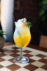 yellow-orange tropical cocktail served in a tall, stemmed hurricane glass, topped with whipped cream or froth and garnished with a mint leaf, set on a distinctive checkered chess board table.