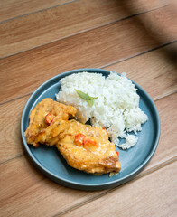 Two pieces of savory chicken cutlet or fried fish smothered in a creamy, chili-spotted sauce, served alongside a mound of fluffy white rice on a round blue plate on a wooden surface.