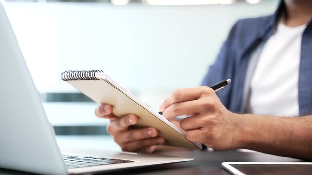 Close up of businessman's hands taking notes in notebook while using laptop at work desk at workplace in office. Worker writes with pen during remote business meeting, conference, seminar or training