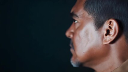 Side profile of a man with short hair and stubble, wearing a brown shirt, gazing to the left in a dimly lit setting. - Powered by Adobe