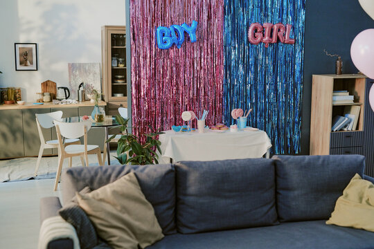 Living room decorated for gender reveal party with pink and blue metallic streamers, balloons, table set with sweets and drinks, kitchen area visible in background, empty sofa in foreground