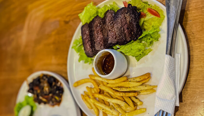 delicious and tempting side view A plate of perfectly grilled steak served on a wooden table with crispy fries with fresh salad and black sauce