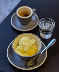 front view of sweet, delicious and tempting Affogato classic: yellow vanilla ice cream with a shot of espresso in a porcelain cup and a glass of water in a small, transparent glass on a black table.