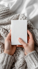 Hands holding blank white book on cozy knitted blanket.  
