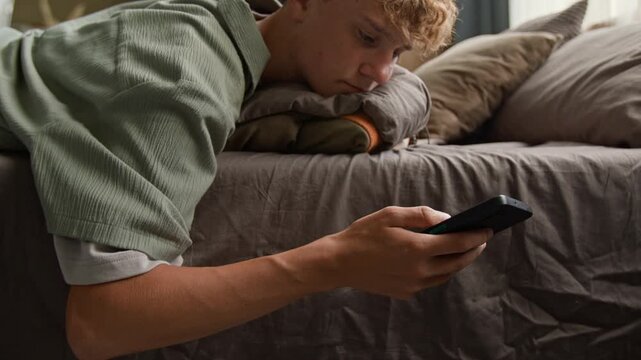 Side view closeup shot of bored young man using smartphone and mindlessly scrolling social media while lying on bed with hand hanging down, copy space