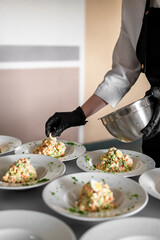 Chef plating traditional Russian Olivier salad on white plates, wearing gloves and apron, preparing for food service or event in professional kitchen setting