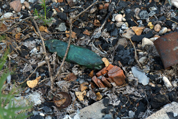 A green glass bottle and broken terracotta among debris and charred remains on a bed of dry leaves, rocks, and dirt.