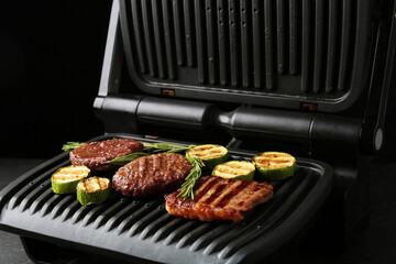Electric grill with beef steaks, zucchini and rosemary on black table, closeup