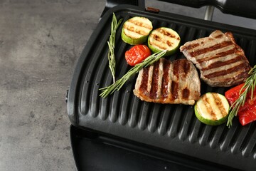 Electric grill with beef steaks, vegetables and rosemary on grey table, closeup