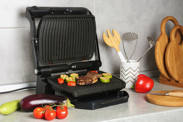 Open electric grill with beef steak, vegetables and cookware on light grey table