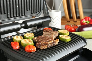 Open electric grill with beef steak, zucchini and tomatoes on table, closeup