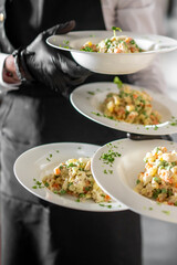 Waiter in black apron holding four plates of creamy vegetable salad with herbs. Professional food service presentation at restaurant or catering event.