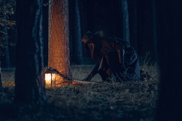 Mysterious figure in dark cloak crouches near lantern amidst shadowy forest trees.