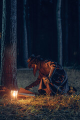 Mysterious plague doctor kneels beside lantern amidst shadowy forest trees.