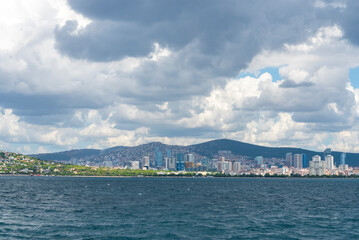 City skyline with mountains in the background and clouds reflecting over the water during midday in a coastal area