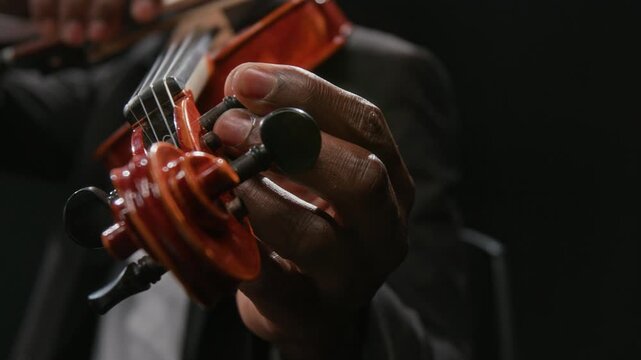 Close up on hand of young adult Black musician tuning violin by adjusting pegs while preparing string instrument for music performance on stage against black background