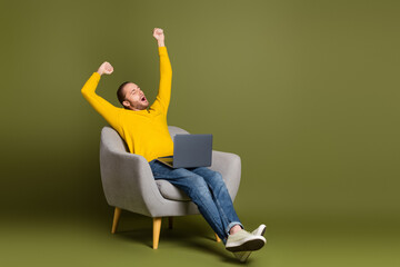 Young man in a casual yellow sweater relaxing in a chair with a laptop on a khaki-colored minimalist background setting.