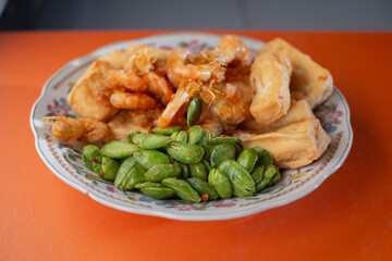 A plate of delicious fried tofu mixed with fried shrimp and stink bean on an orange table