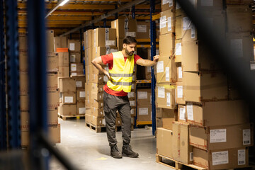 A focused warehouse worker checks inventory while surrounded by stacks of cardboard boxes in a well-organized storage area.