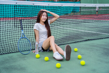 Young woman sitting on tennis court with racket and balls, looking relaxed and happy