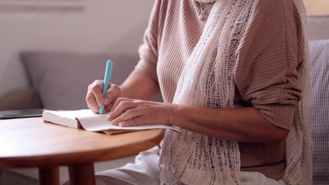 Older Asian woman writing notebook at home comfortable setting daily life planning thoughts senior citizen learning journaling