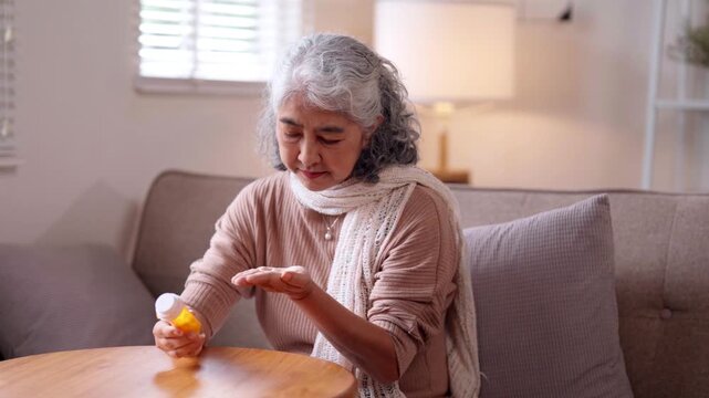 Older Asian woman carefully taking daily medication at home managing health senior wellness routine self care elderly life comfort indoors apartment