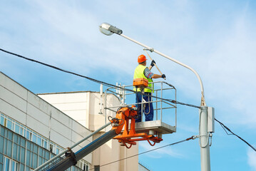 Painter in lift bucket paint street lighting pole at height. Man in cradle painting lamppost. Utility worker paint street light pole, painting lamppost with brush. Worker renovate lightning pole © Tricky Shark