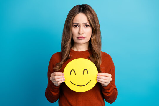 Charming woman holding a yellow smiley face cutout while appearing sad against a bright blue background