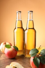 Delicious cider in glass bottles and apples on wooden table against orange gradient background