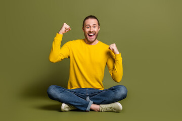 Young cheerful man sitting cross-legged excitedly celebrating with fists raised wearing bright...