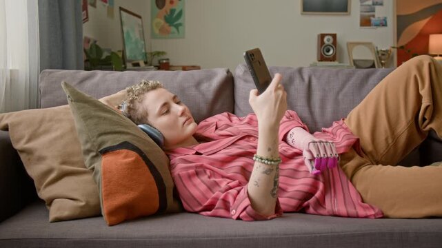 Young woman with prosthetic hand using smartphone and scrolling social media while lying on couch at home and relaxing