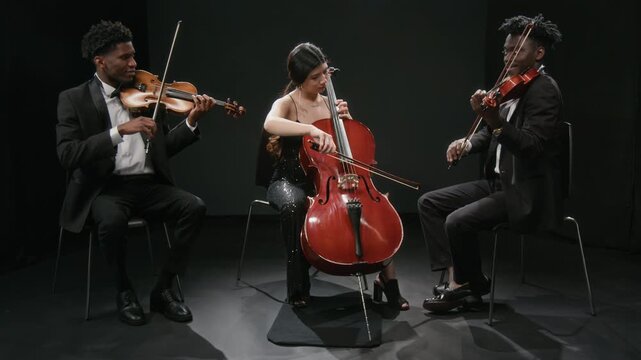 Full length shot of diverse group of three young adult symphony orchestra musicians playing violins and cello while performing music on dimly lit stage against black curtains