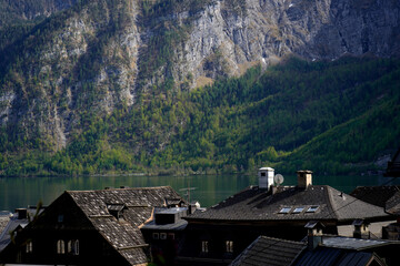 Layered rooftops with shingles, chimneys and skylights face a still lake below steep forested...