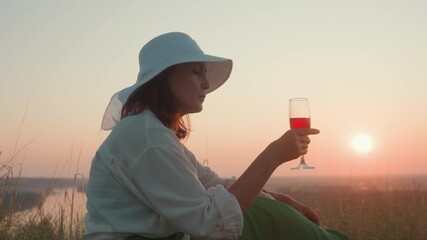 Profile view of woman wearing wide brim hat sipping red wine from glass during sunset picnic in grassy field overlooking river and horizon, soft warm light creating relaxed outdoor atmosphere - Powered by Adobe