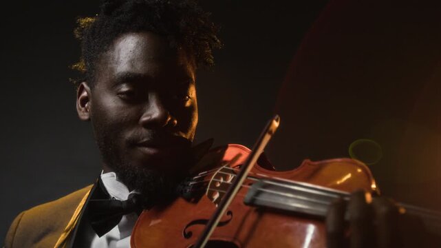 Concentrated African American male musician wearing bow tie playing violin during music concert on stage against spotlight and dark background