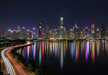 Fototapeta premium Vibrant city skyline at night with dazzling lights reflecting on calm water and light trails on highway