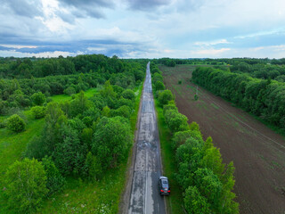 A low-angle aerial view of a black SUV driving on a long, wet, rural road, with a ploughed field on one side and a forest on the other.