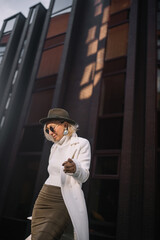 Stylish woman in a white coat and hat poses confidently outdoors against a modern urban backdrop