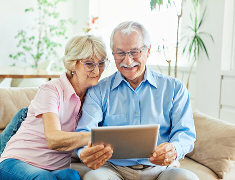 Portrait of a lovely senior mature couple together holding a tablet and having video call sitting on sofa at home