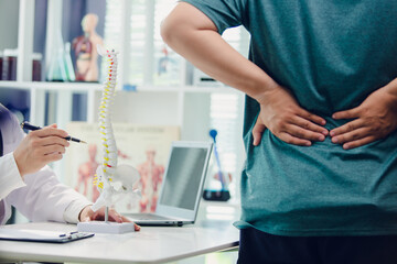 A doctor is giving advice to a patient with back pain in a clinic using a skeleton model, laptop and whiteboard to record the patient's symptoms. Concept of healthcare and medical care.