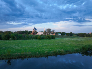 An aerial view of the Suzdal Kremlin with its white walls and blue domes, located behind a lush green meadow and a small river at sunset.