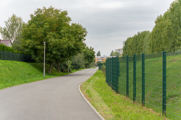An asphalt path leads along a green fence. Trees and houses stand to the right and left.