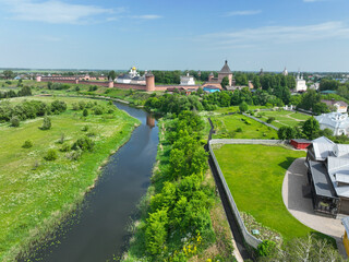 Fototapeta premium An aerial view of a large monastery complex with red brick walls and towers, next to a small river and green fields.