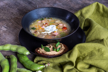 Fresh vegetable soup served in a black bowl with a dollop of cream, accompanied by a slice of bread spread with hummus and green beans on a rustic wooden table