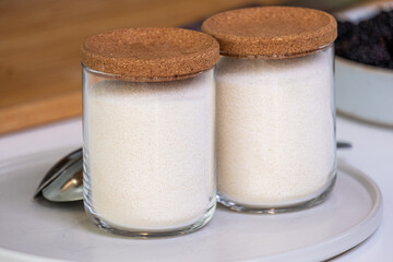 Two glass jars filled with white sugar, topped with cork lids, placed on a white plate beside a silver spoon, showcasing a clean and organized kitchen setting